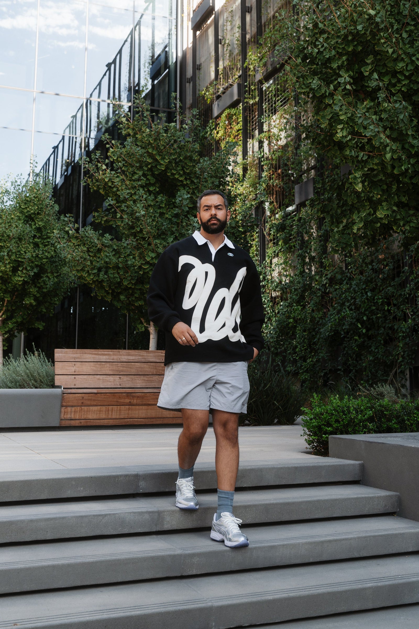 Man standing on steps outdoors wearing a black hoodie with white text, gray shorts, and white sneakers.
